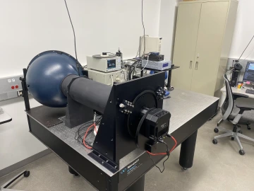 Optical table with equipment used to characterize sensors, including integrating sphere, monochrometer, plasma light source, and temperature controller. A camera is hooked up to baffle leading to integrating sphere.